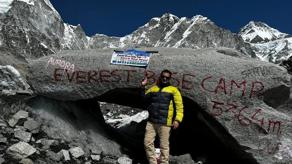 Hiker at Everest Base Camp holding Simrik Real Nepal Treks flag, surrounded by towering Himalayan mountains