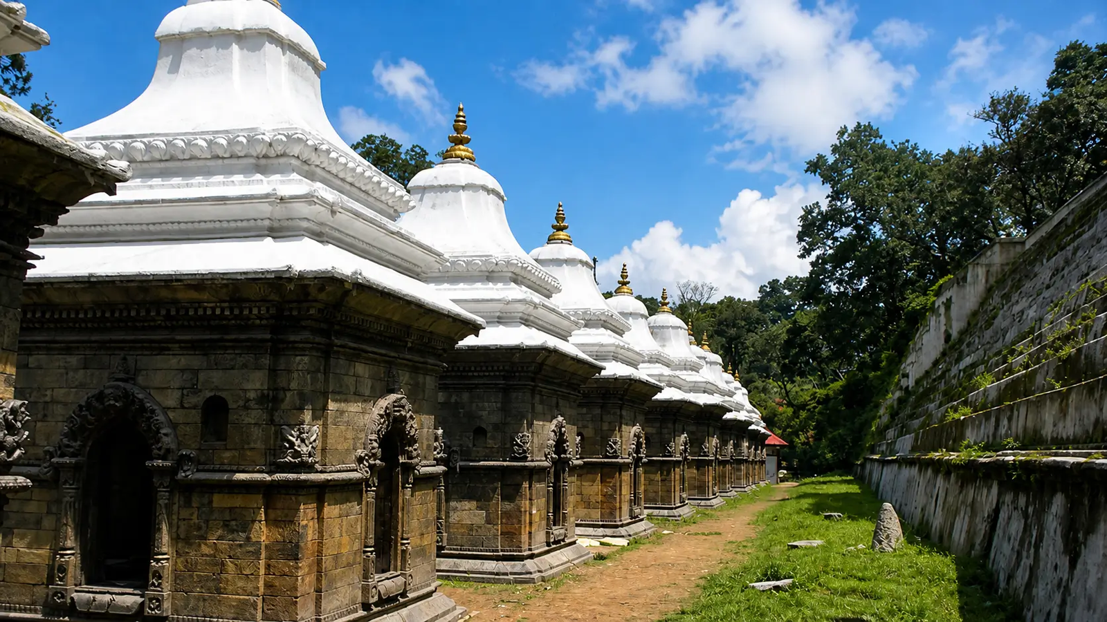 Row of sacred Hindu shrines at Pashupatinath Temple with white domes and intricate stone carvings under blue sky, UNESCO World Heritage site in Kathmandu Nepal