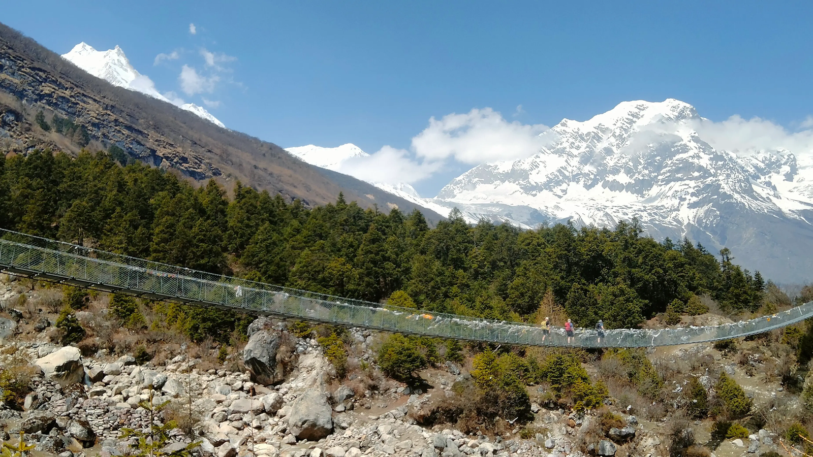 Trekkers crossing a suspension bridge on the Manaslu Circuit Trek route from Syala to Samagaon, with the Budhi Gandaki River valley and Himalayan mountain scenery in Nepal.
