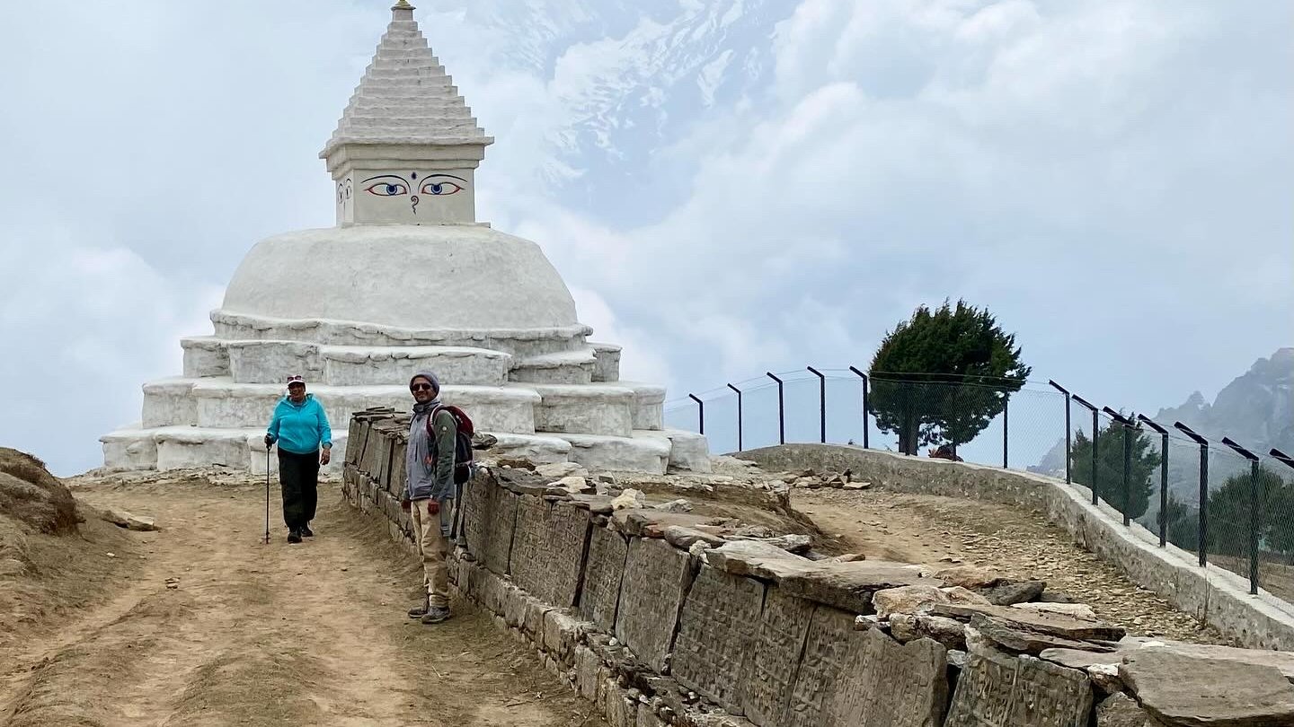 Trekking near a white Buddhist stupa and mani wall on the Everest View Trek route in the Khumbu region of Nepal, with snow-covered mountains hidden in clouds.