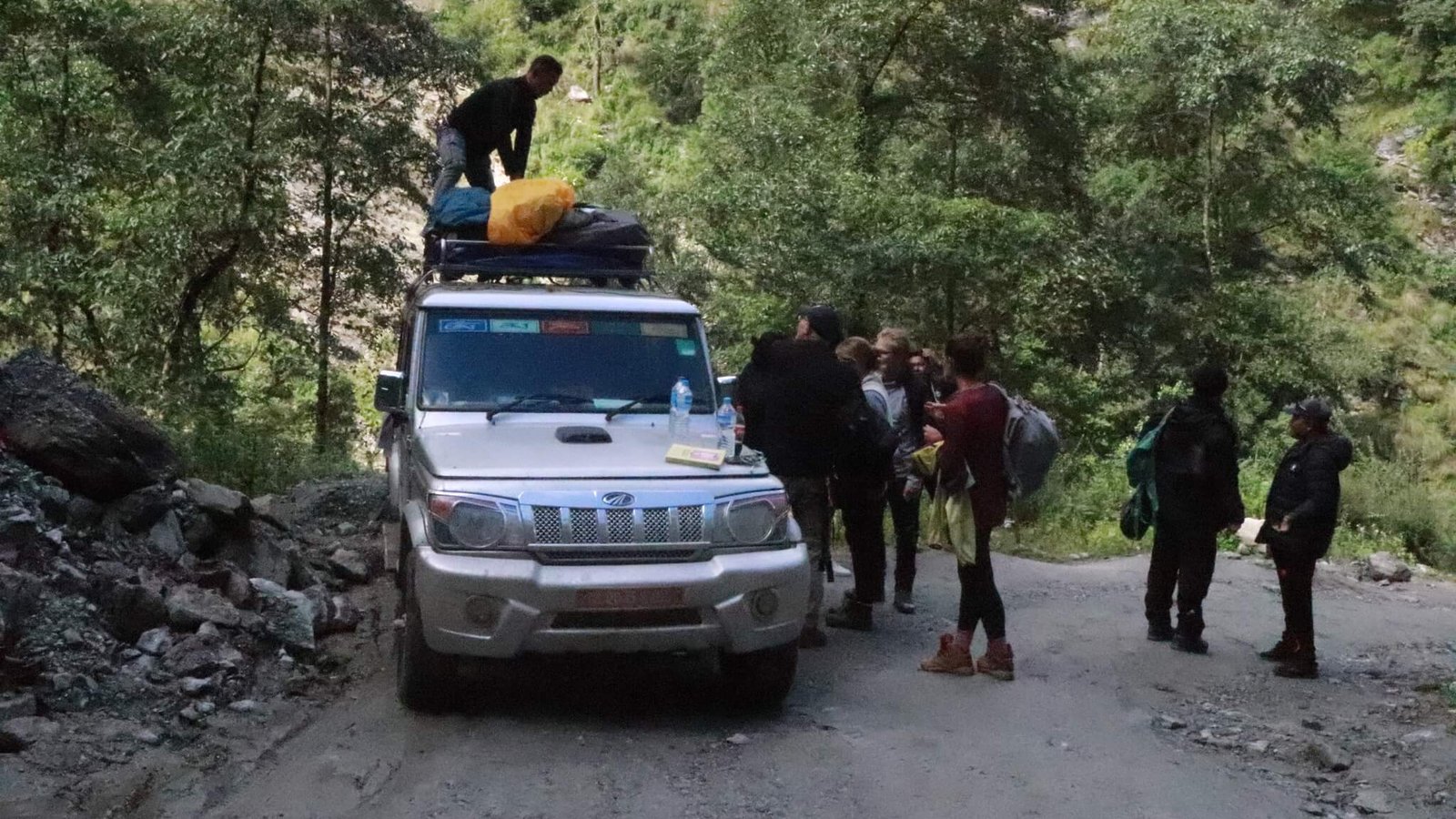 Overland journey to Surke as an alternative to Lukla flights for Everest Base Camp trek, showing travelers preparing luggage on a jeep on a mountainous dirt road surrounded by forest.